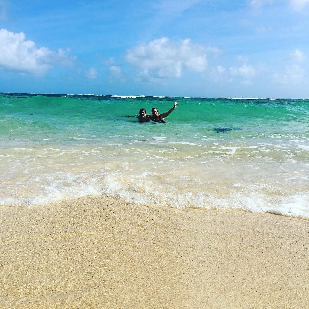 Deux personnes sur une plage dans l'océan turquoise, l'une d'elles levant le bras. Plage de sable, ciel bleu avec nuages.