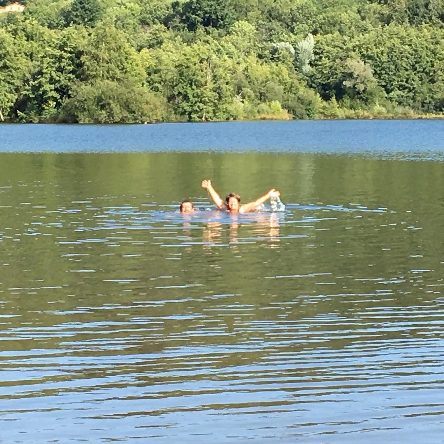 Deux personnes nageant dans un lac, les bras levés et souriant. Des arbres verts bordent la rive sous un ciel bleu.