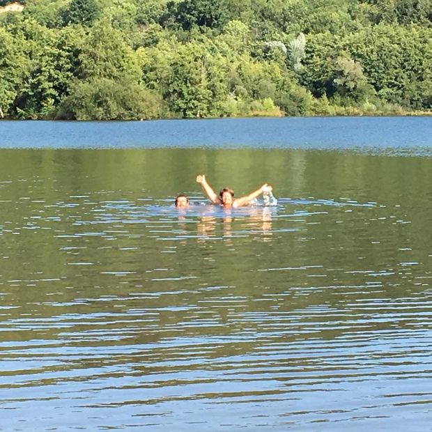 Deux personnes nageant dans un lac, les bras levés et souriant. Des arbres verts bordent la rive sous un ciel bleu.