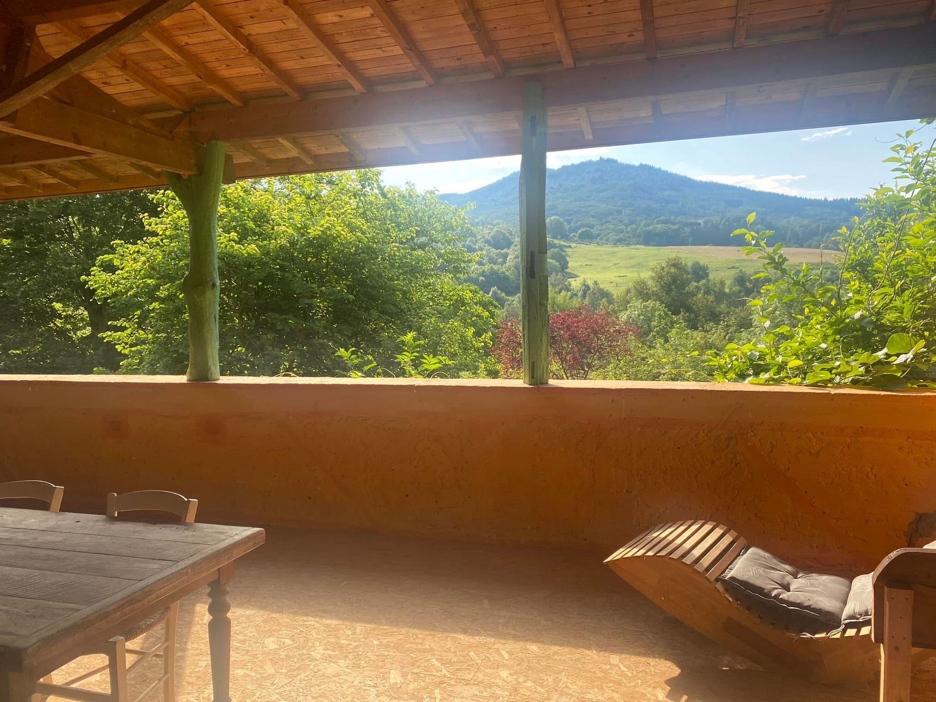 Véranda donnant sur un paysage verdoyant avec une montagne au loin. Une table en bois et une chaise longue sont installées.
