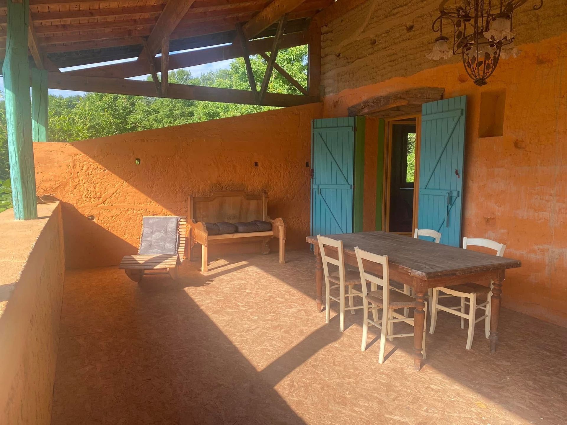 Terrasse extérieure aux murs en stuc orange, volets bleus et table en bois avec chaises blanches.