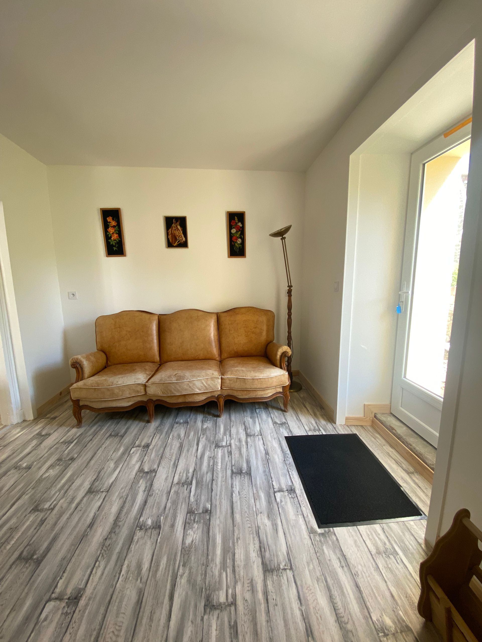 Un salon avec un canapé jaune usé, trois cadres et une porte baignée de lumière. Parquet et paillasson foncé.