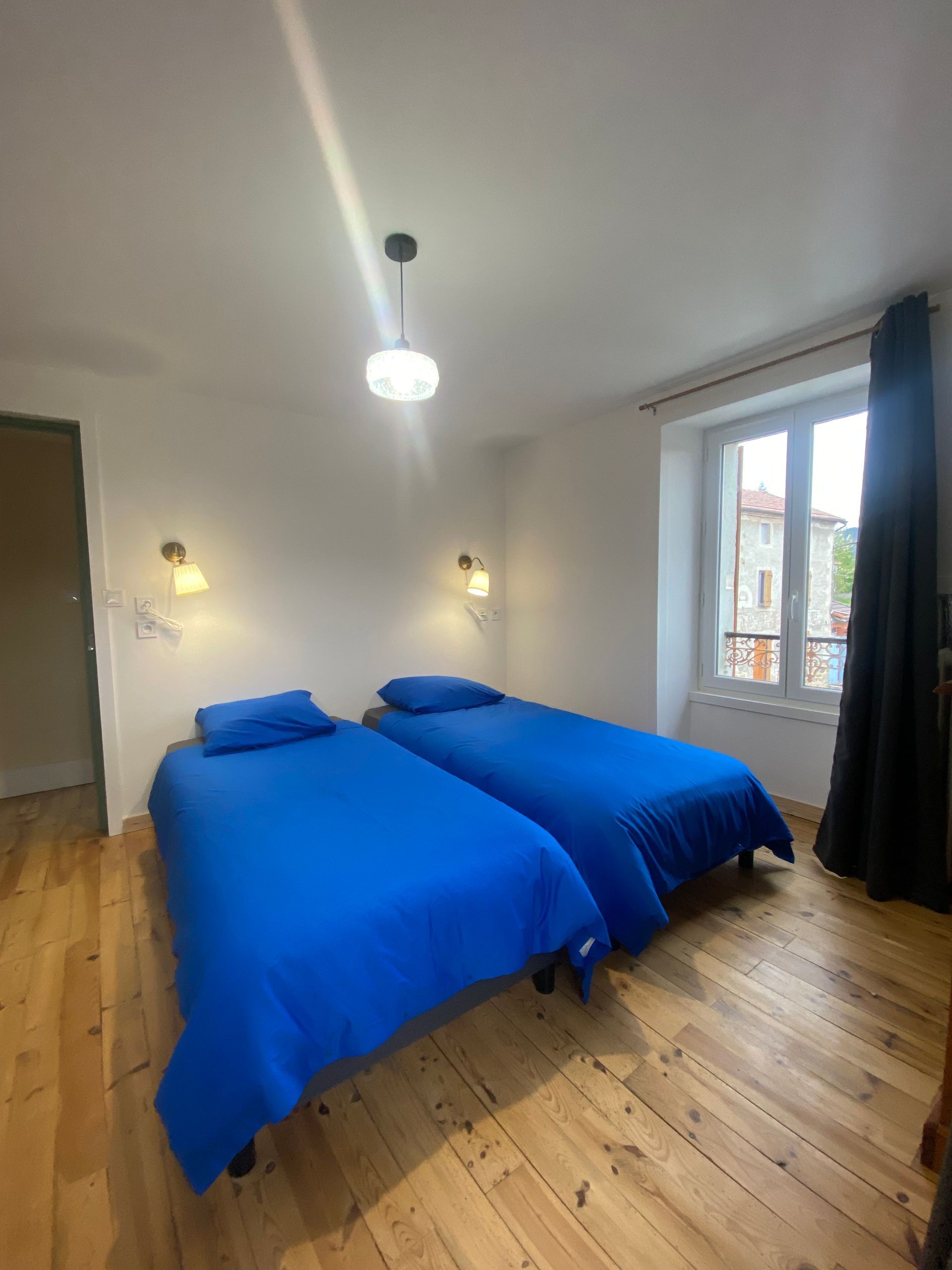 Deux lits jumeaux avec literie bleue dans une chambre avec parquet et une fenêtre avec rideaux noirs.