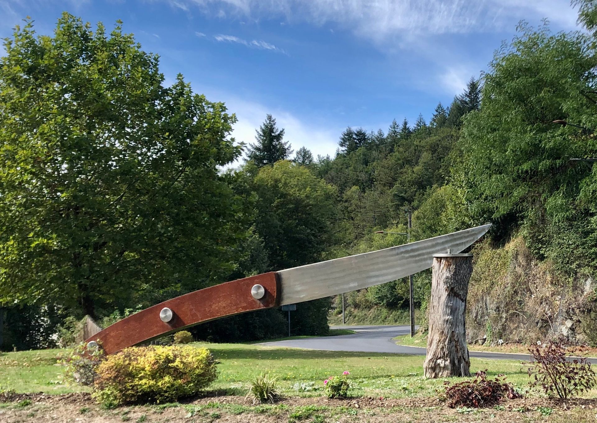 Une grande sculpture d'un couteau, avec un manche brun rouille, une lame en métal et reposant sur une souche d'arbre.