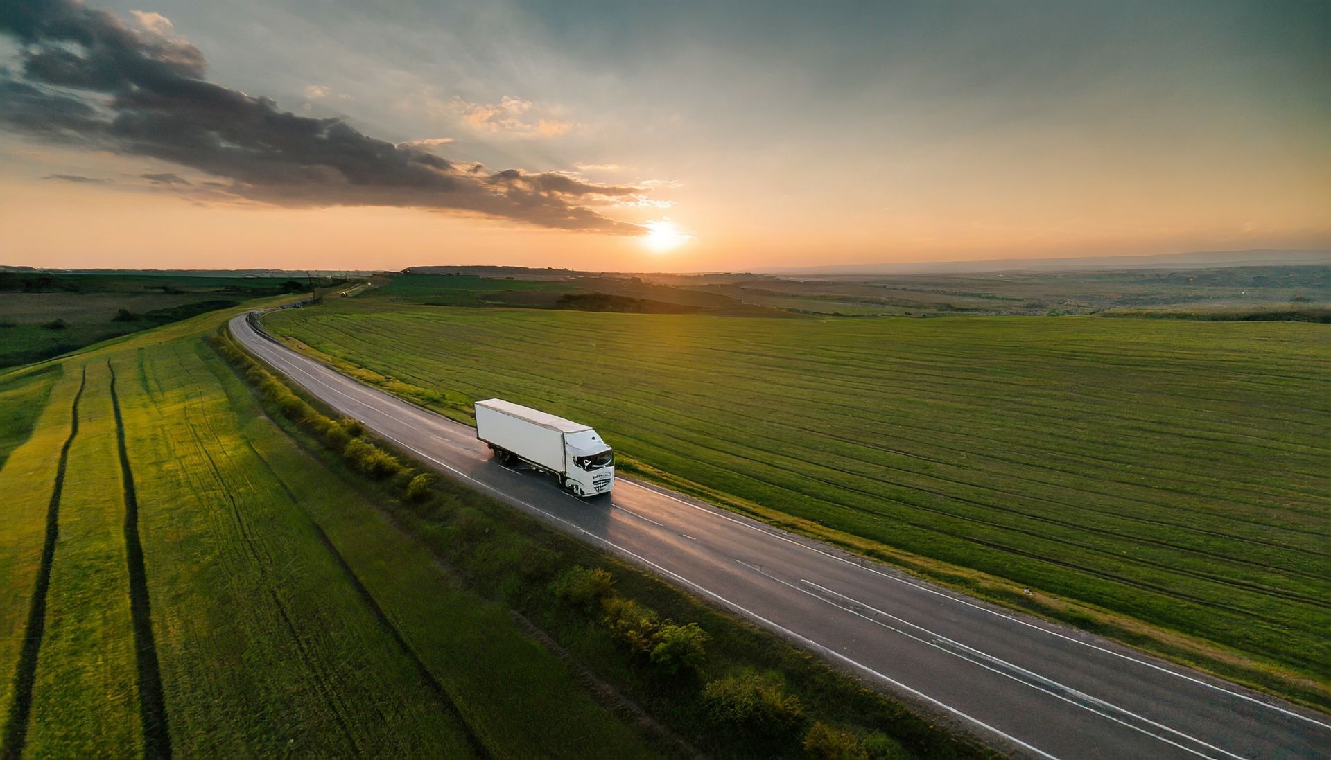 An aerial view of a semi truck driving down a country road at sunset.