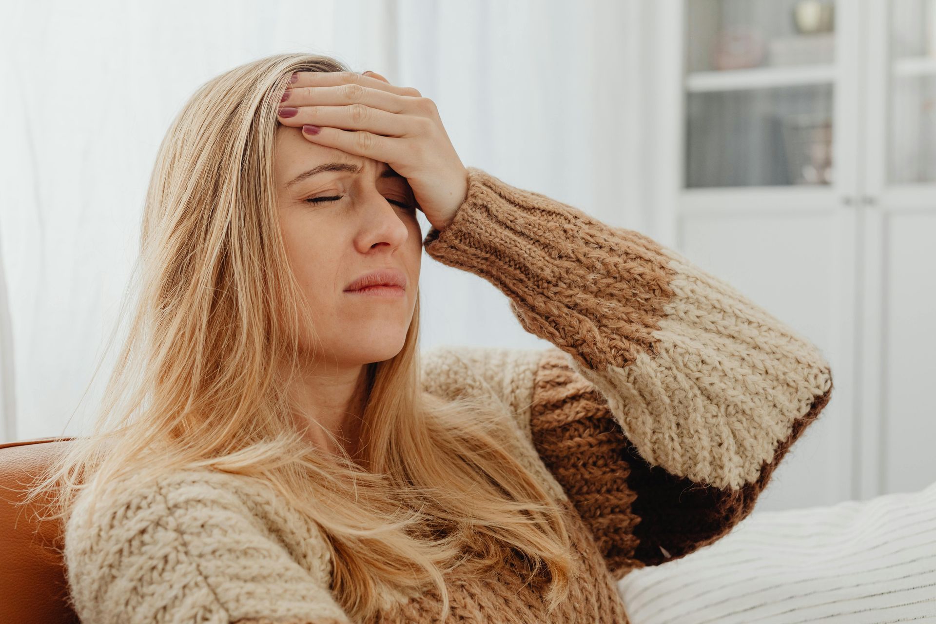 A woman sits on a couch, rubbing her palms against her forehead, clearly stressed or overwhelmed.