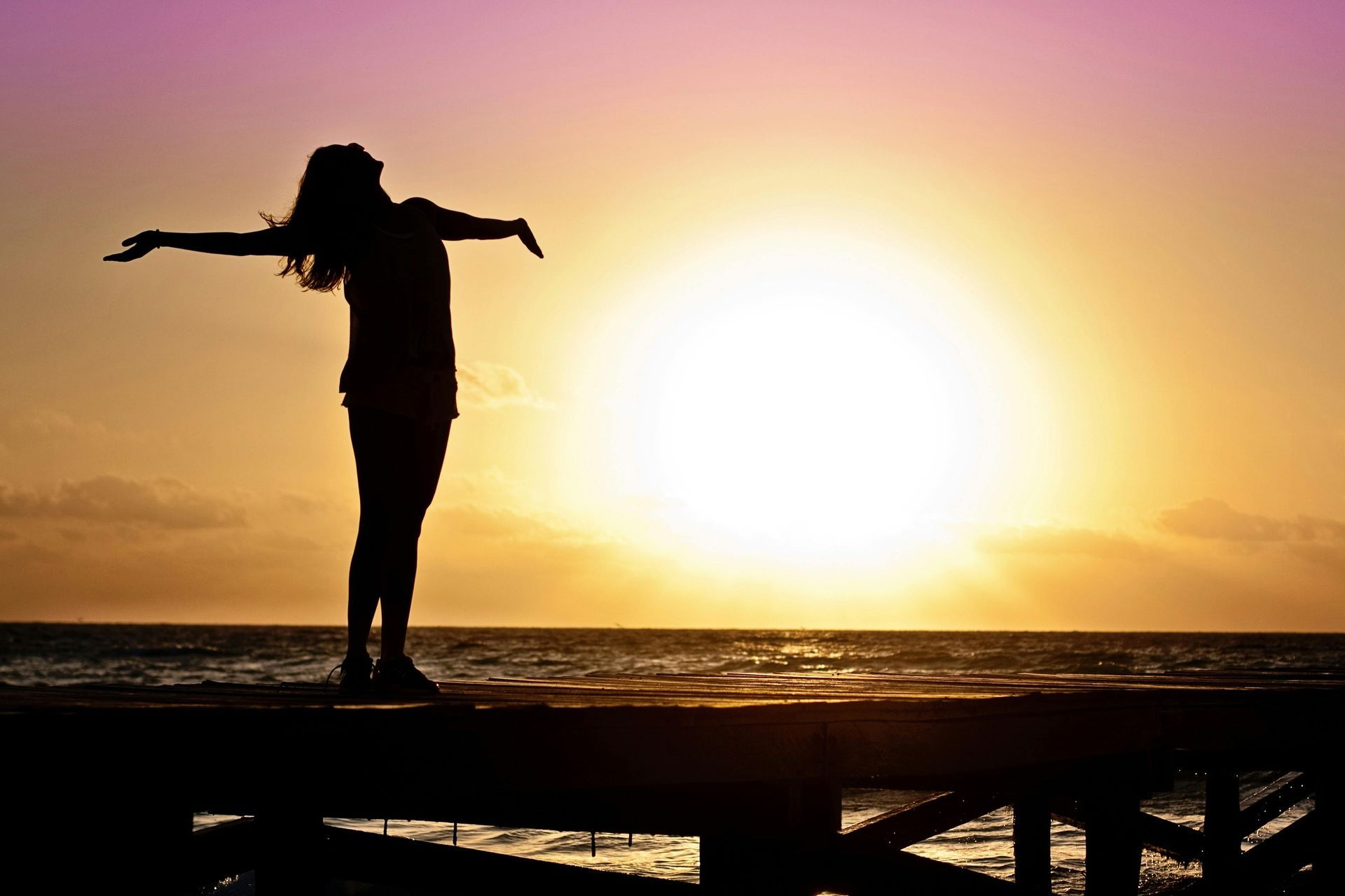A silhouette of a woman standing on a bridge with her arms wide open with sunset in background