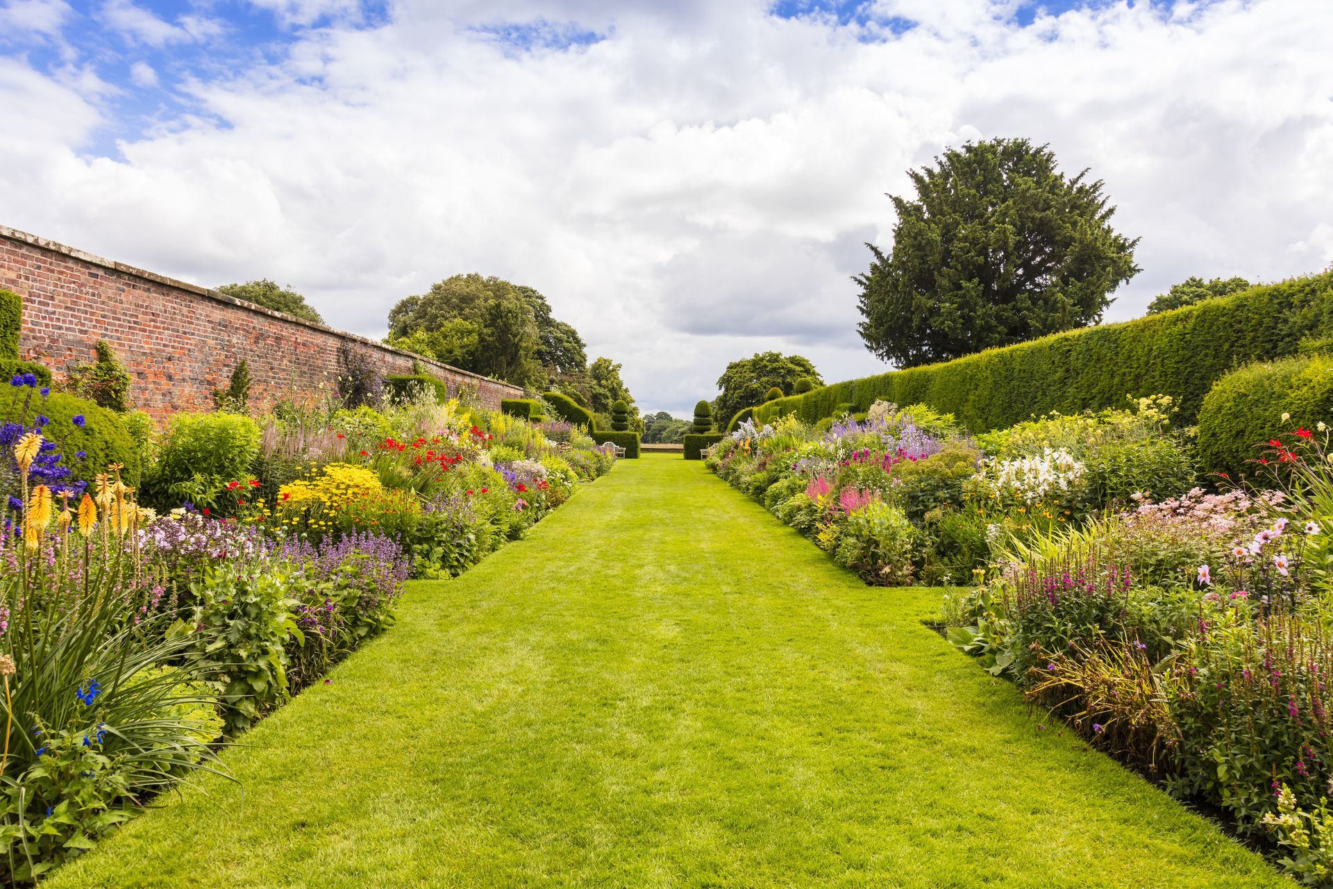 Lush green grass path leads through vibrant flowerbeds in a garden, under a cloudy sky.