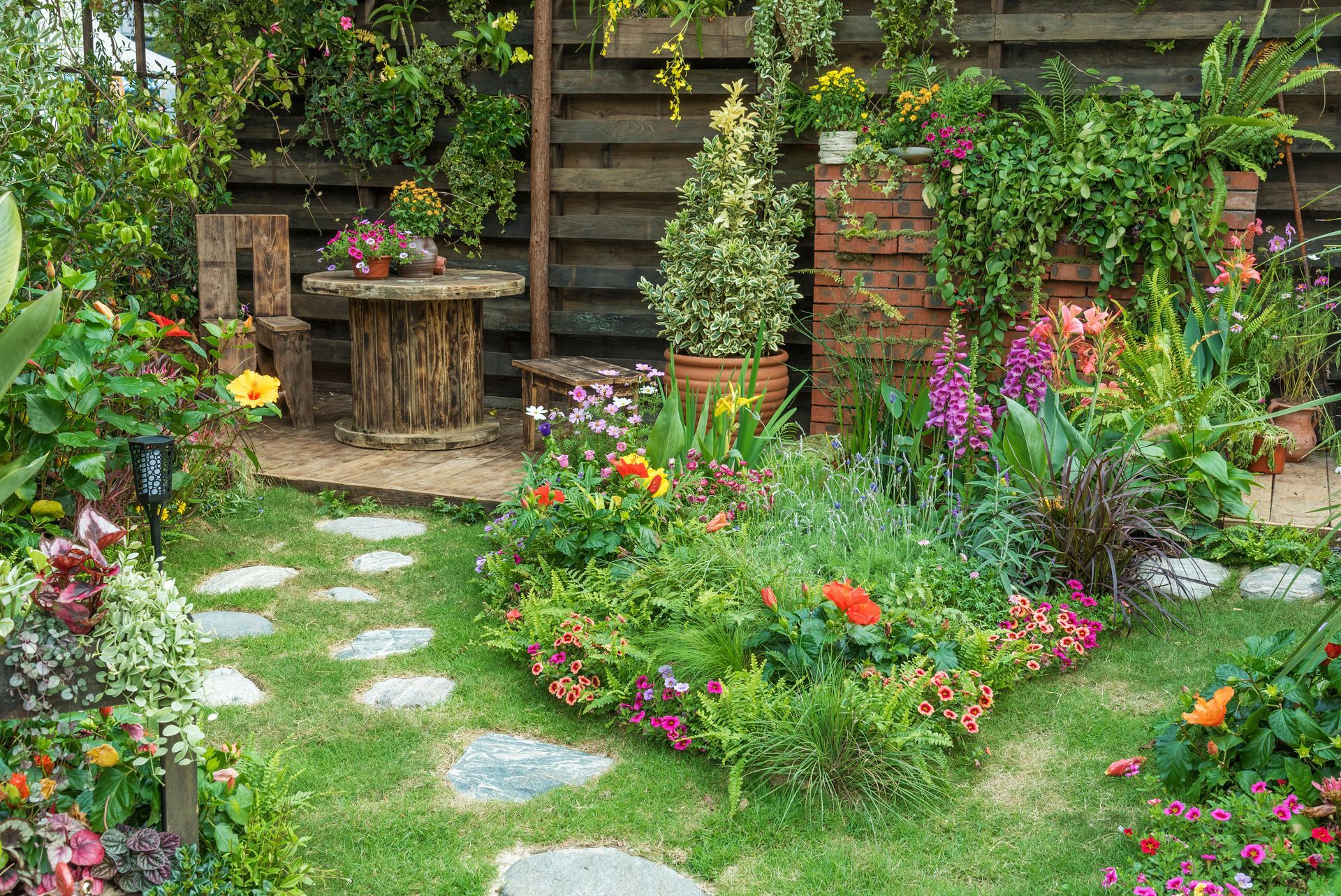 A colorful, lush garden with a stone path, wooden table and chair.
