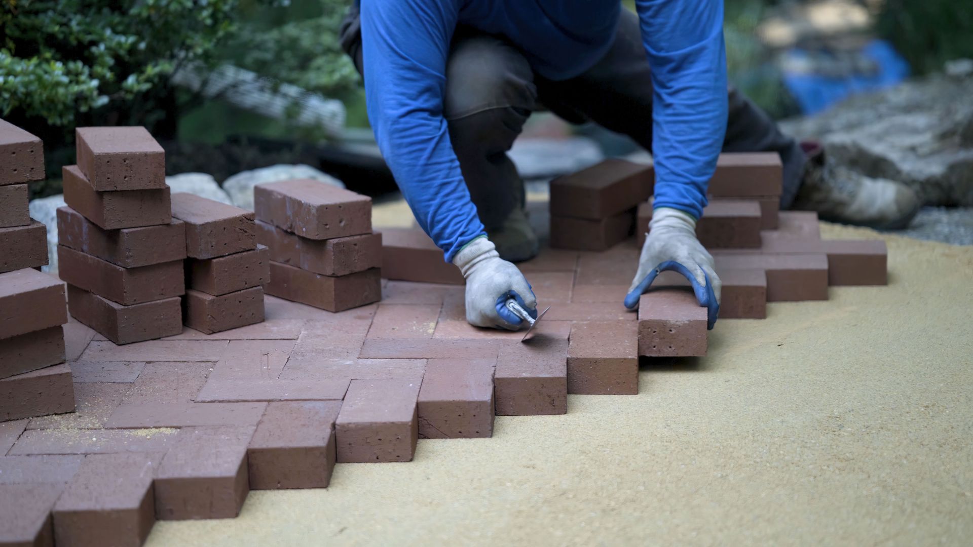 Person laying bricks in a herringbone pattern on a sand base, wearing gloves and a blue shirt.