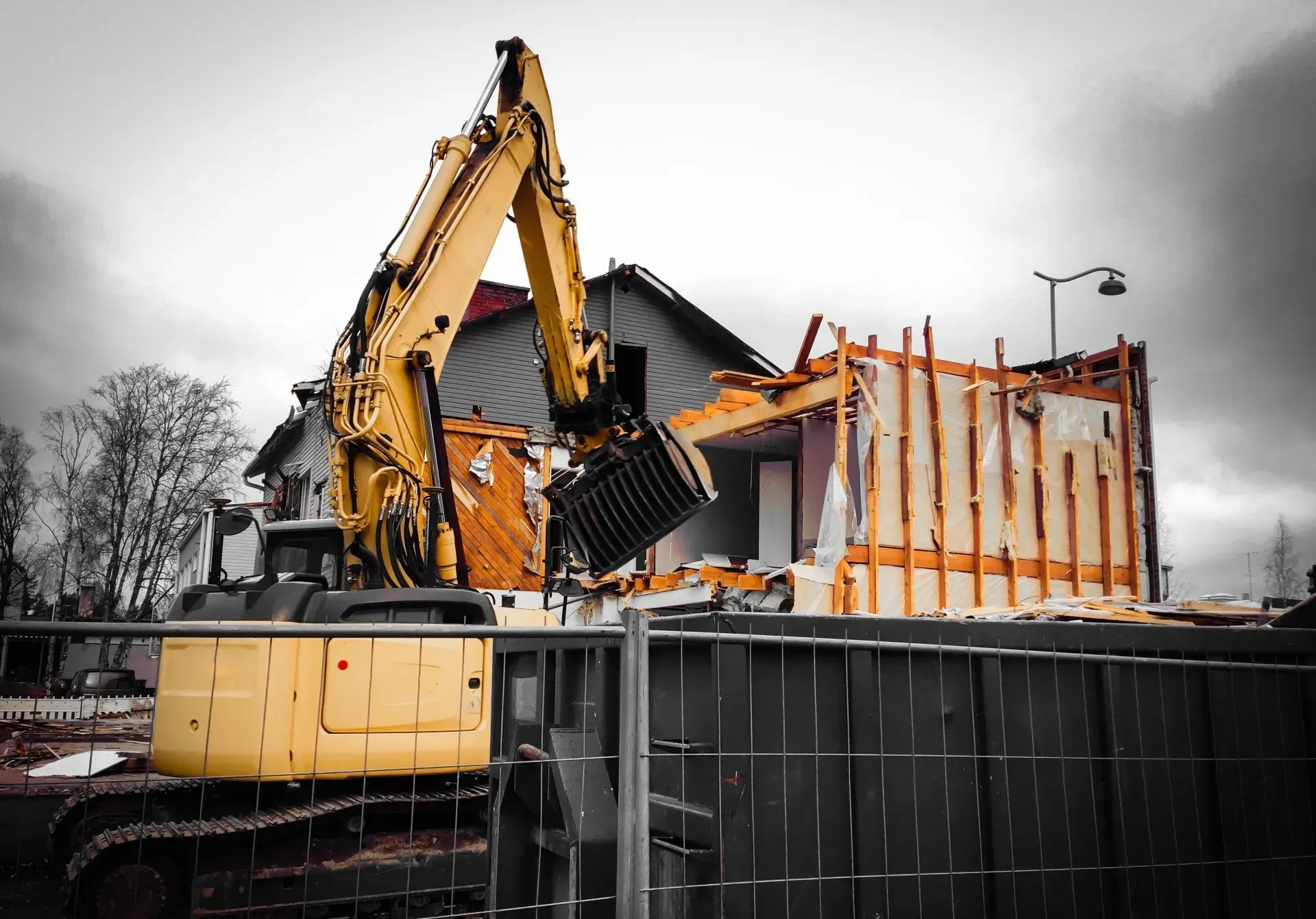 Yellow excavator demolishing a building on a cloudy day, behind a black fence.