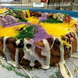 A King Cake decorated with purple, gold, and green sugar and white icing, sitting on a paper-lined tray.