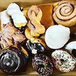 A cardboard box filled with an assortment of donuts, including glazed, chocolate-frosted, cinnamon roll, and sprinkles.