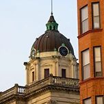 A domed, cream-colored building with clock faces sits next to a tall, brown brick building against a light sky.