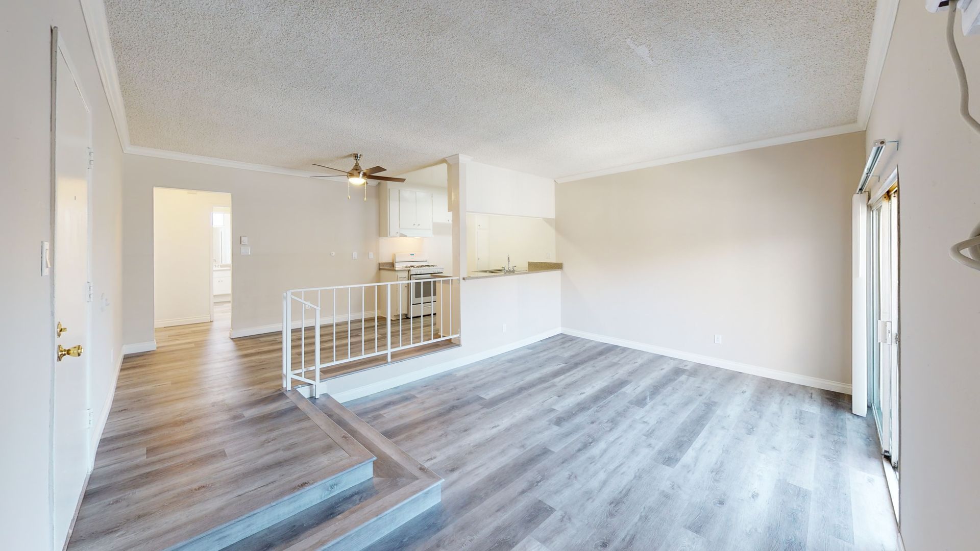 Empty, neutral-toned living space with wood-look flooring, connecting to an entrance and a raised kitchen area with a small railing.