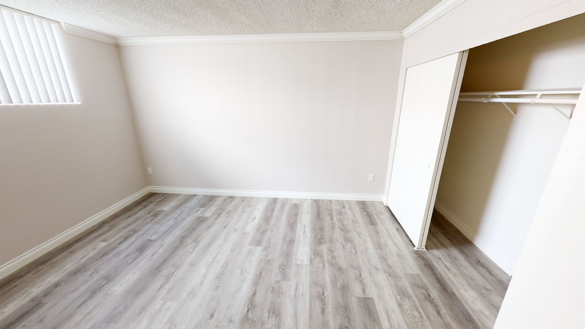 Empty bedroom with light gray wood-look flooring, white walls, closet with a hanging rod, and a window with blinds.