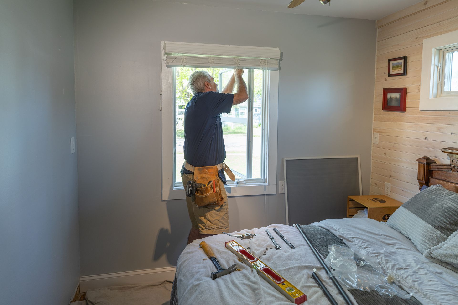 A man is installing blinds on a window in a bedroom.