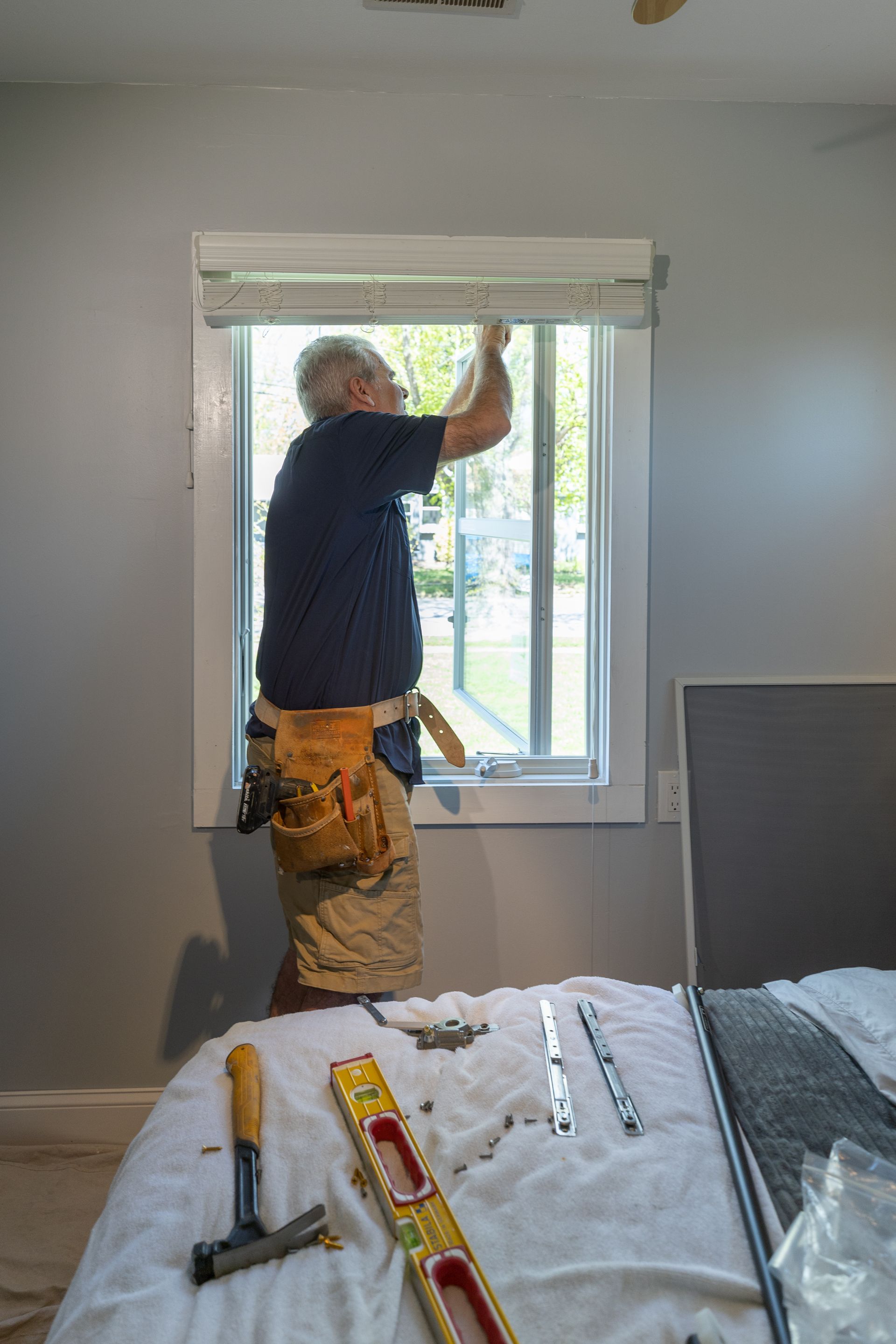 A man is installing blinds on a window in a bedroom.