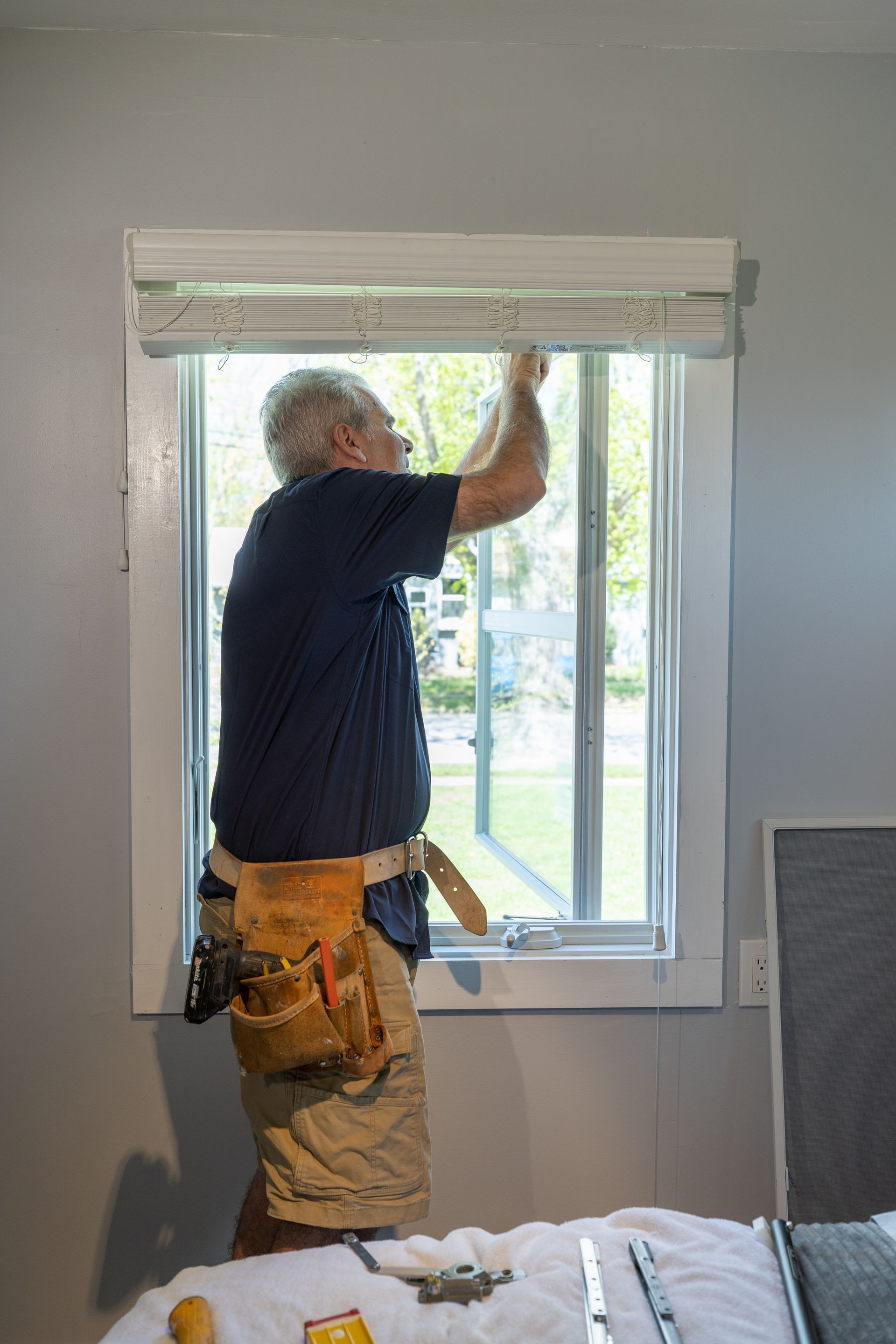 A man is installing blinds on a window in a bedroom.