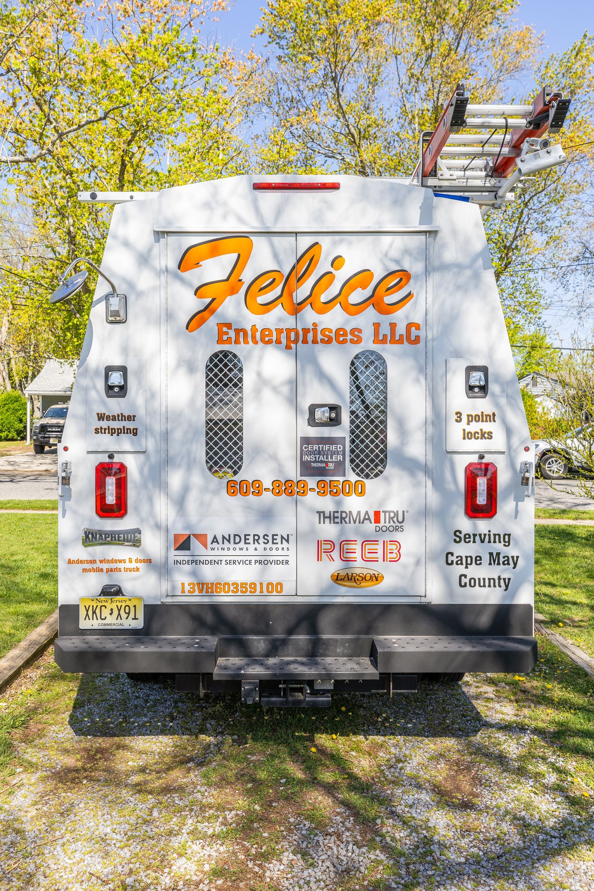 The back of a white van with a ladder on top of it is parked in a grassy area.