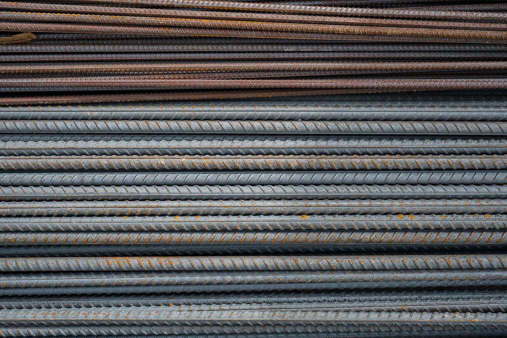 Una pila de barras de acero corrugado paralelas y acanaladas, que van de color marrón oxidado en la parte superior a gris metálico en la inferior.