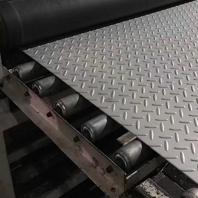 Una lámina de metal gris con patrón de diamante descansa sobre una cinta transportadora con rodillos visibles en un entorno industrial.