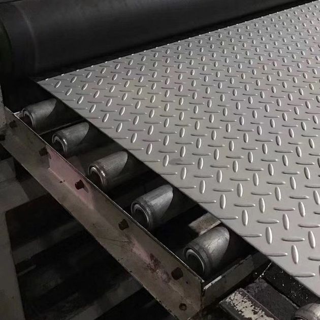 Una lámina de metal gris con patrón de diamante descansa sobre una cinta transportadora con rodillos visibles en un entorno industrial.