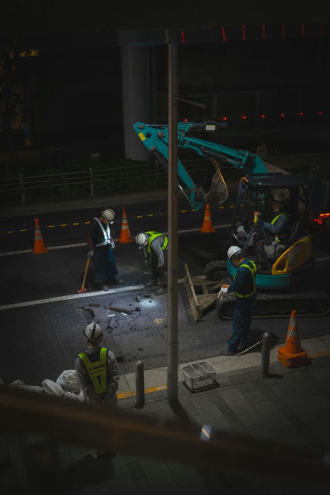 Obreros de la construcción con chalecos reflectantes utilizan una pequeña excavadora para realizar trabajos nocturnos de reparación de calles cerca de conos de tráfico naranjas.