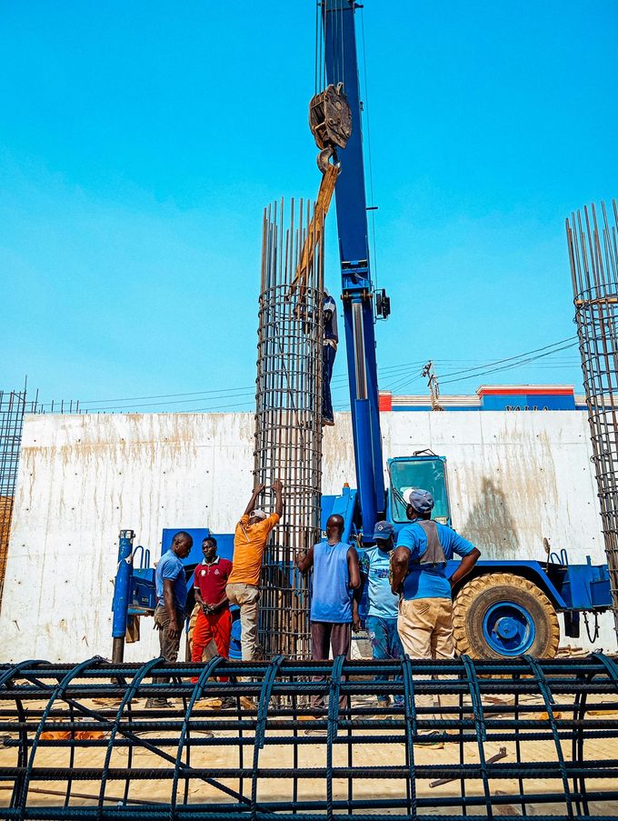 En una obra, unos obreros de la construcción guían una grúa que eleva una jaula cilíndrica alta de barras de refuerzo para un pilar de hormigón.