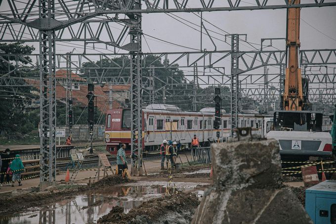 Un tren de cercanías estacionado en una zona de construcción embarrada, bajo líneas eléctricas aéreas, con maquinaria pesada cerca.