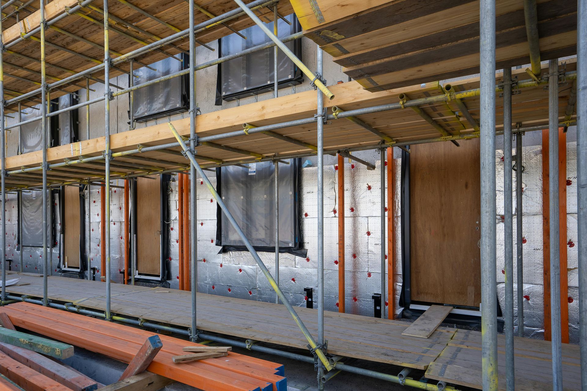 Un andamio rodea un edificio en construcción, dejando al descubierto paneles aislantes metálicos, ventanas tapiadas y pilas de madera.