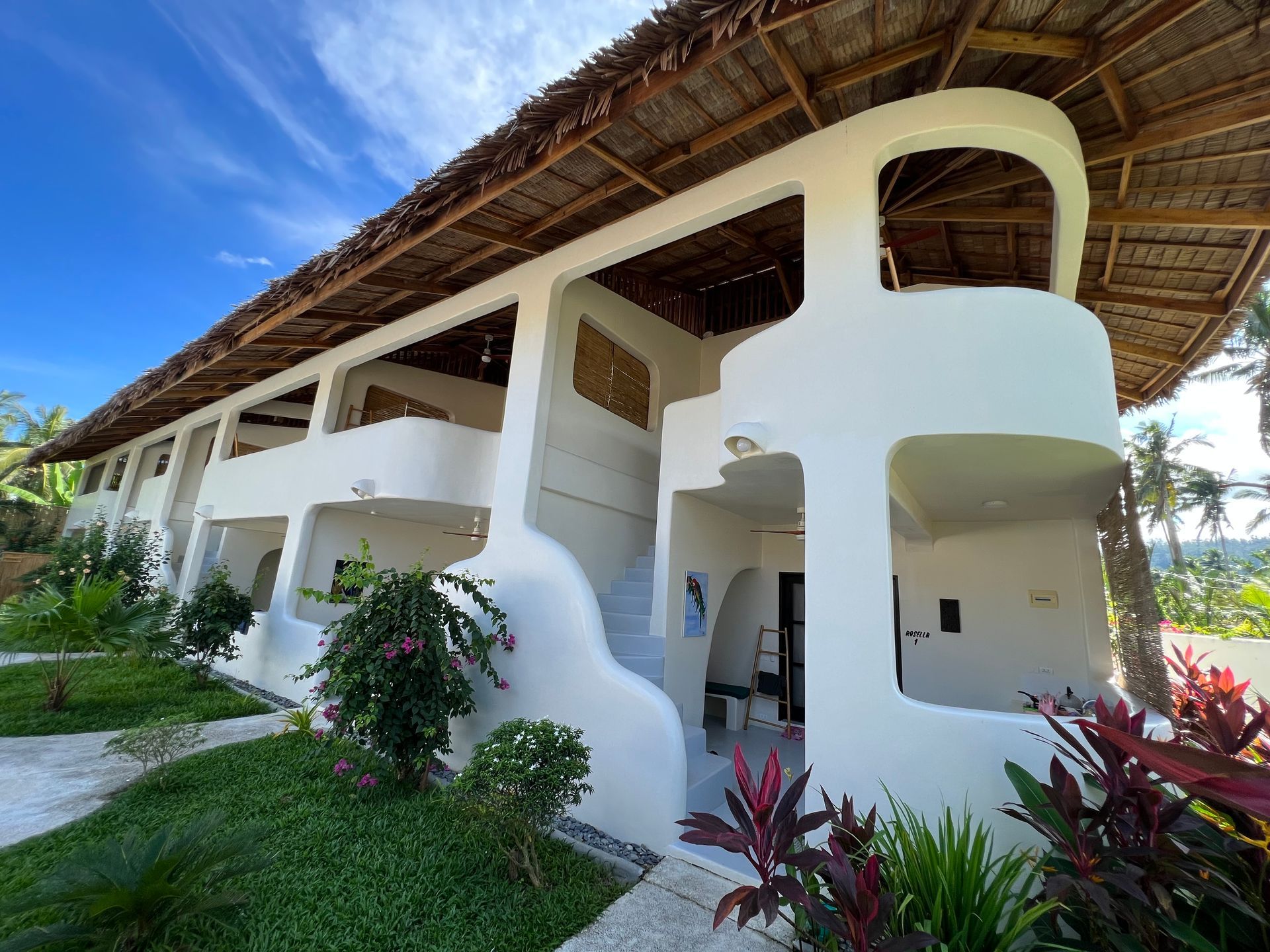 A large white building with a thatched roof is surrounded by plants.