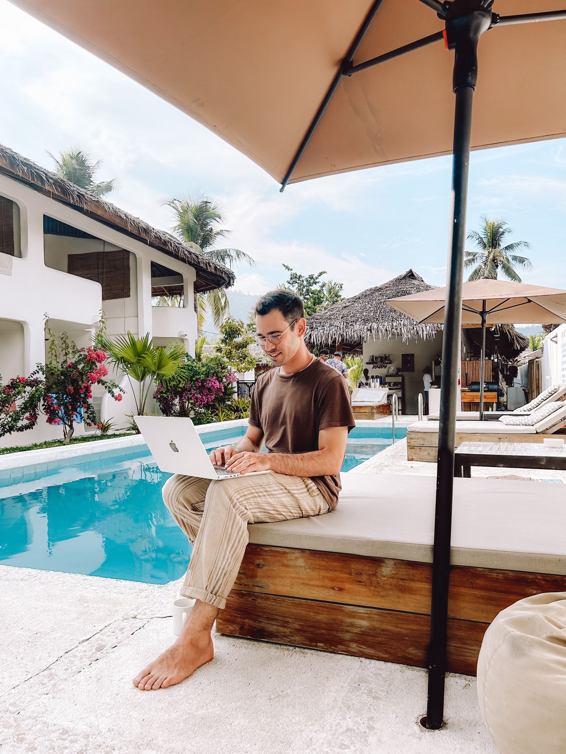 A man is sitting on a bench in front of a swimming pool using a laptop computer.