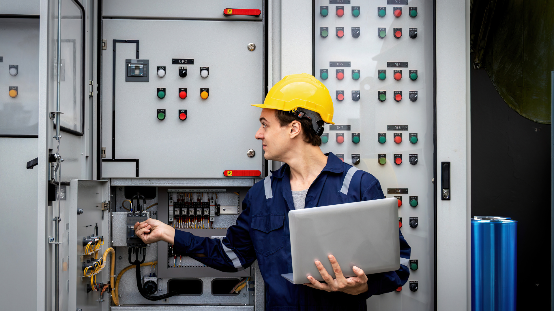 A man in a hard hat is holding a laptop in front of a control panel.