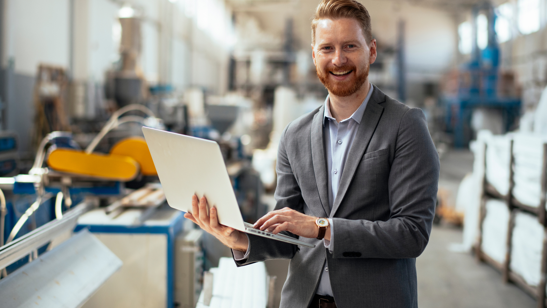 A man in a suit is holding a laptop computer in a factory.