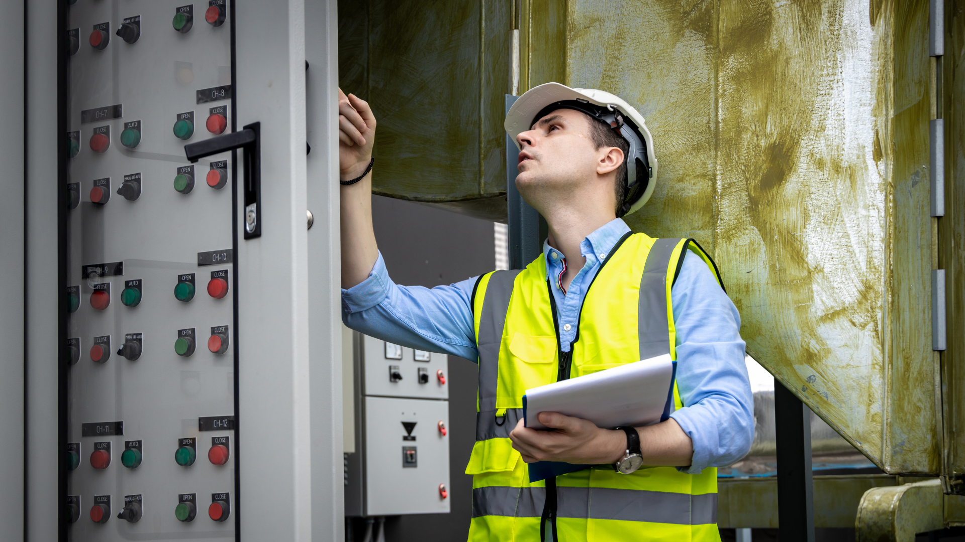 A man in a yellow vest and hard hat is looking at a control panel.