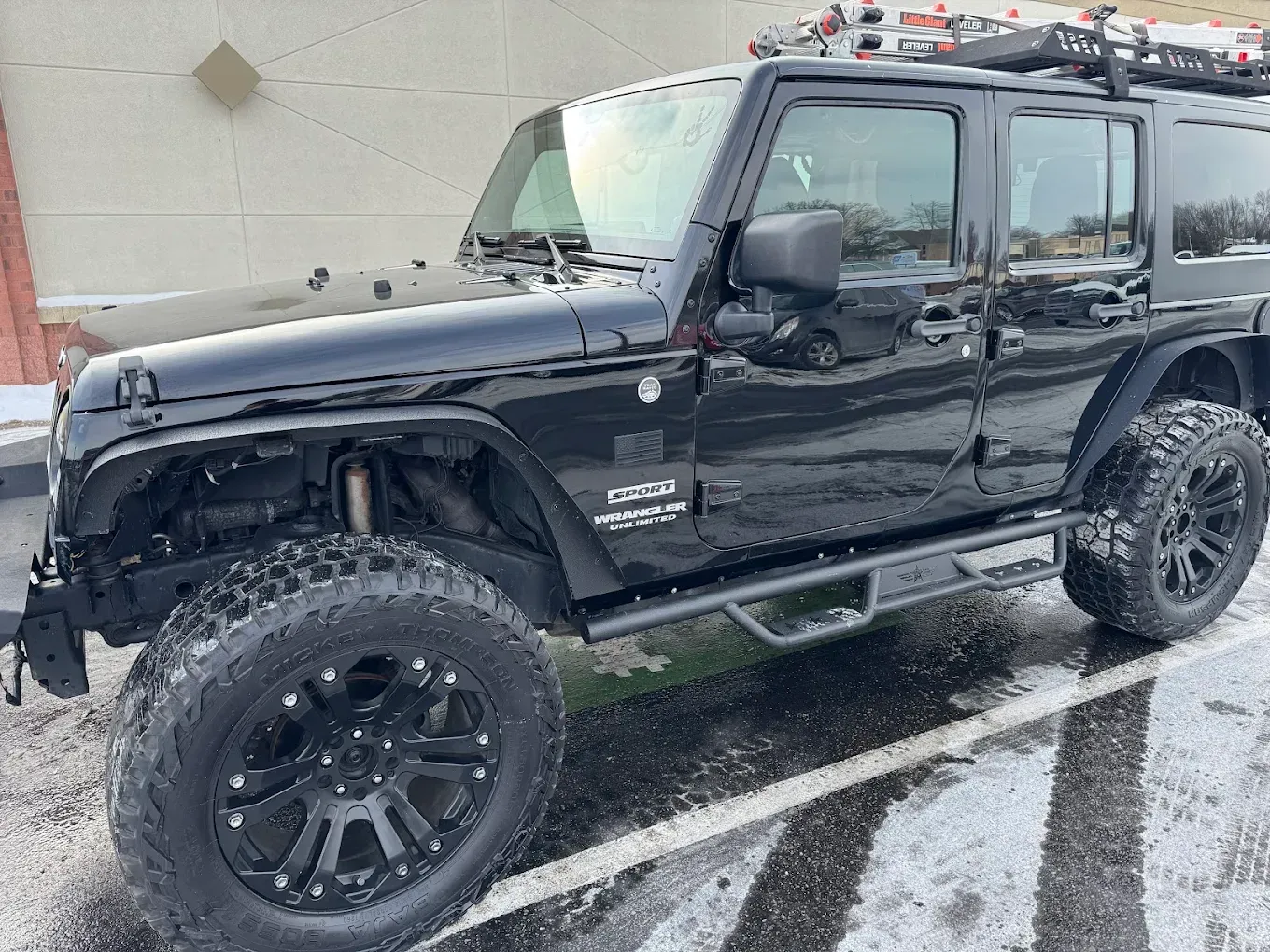 Black Jeep Wrangler parked on a snowy surface, with a roof rack and oversized tires.