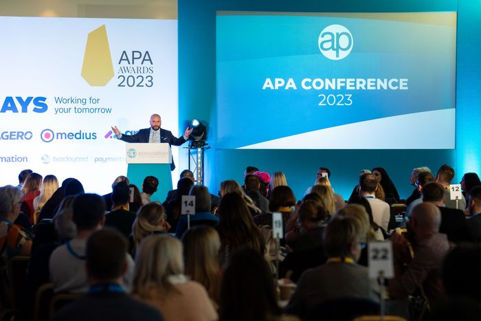 A man is giving a speech in front of a crowd at an apa conference.