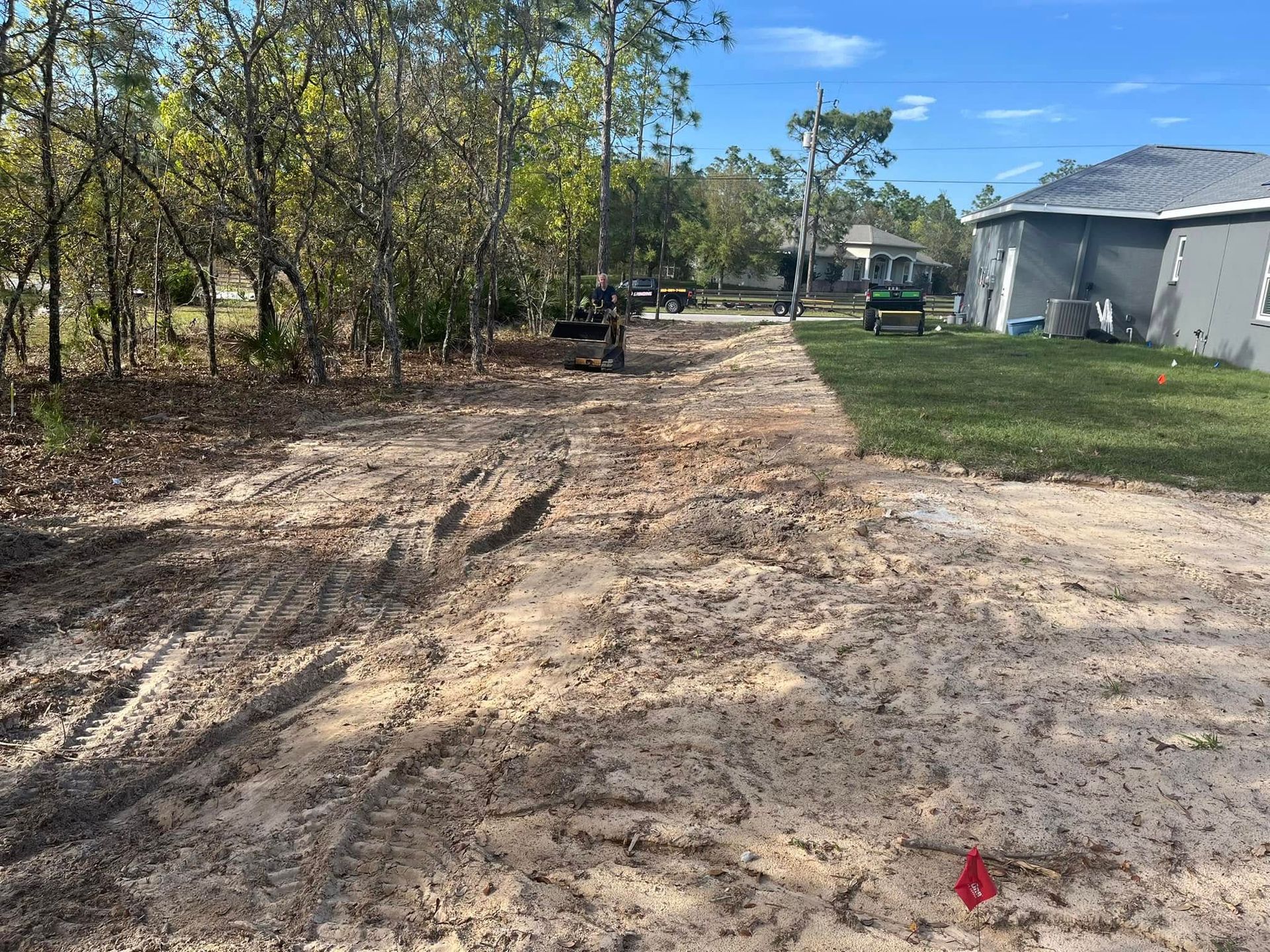 Dirt path next to a grassy area with a house on the right, trees and a small construction vehicle in the distance.