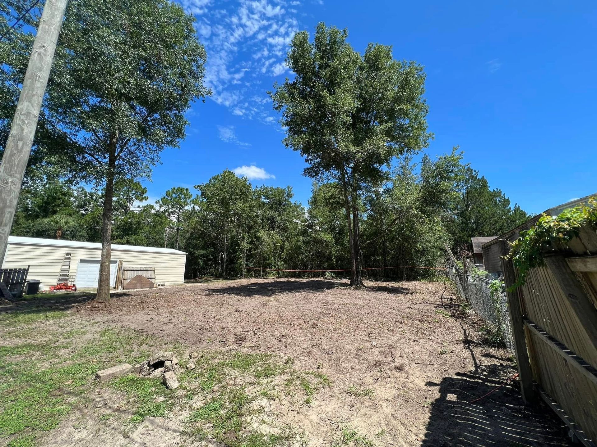 Open lot of dirt with some trees and a blue sky. A building is in the background.