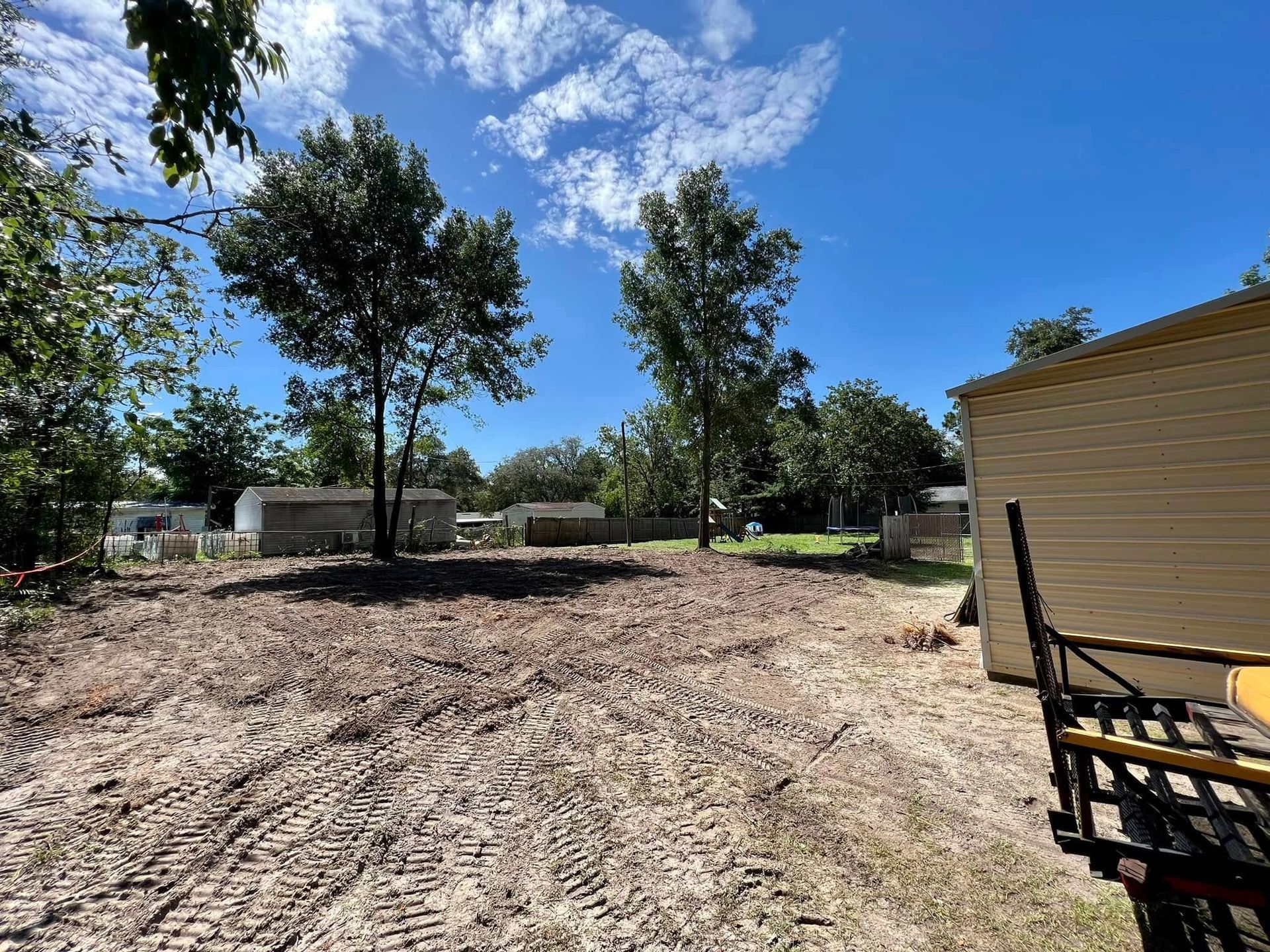 Cleared dirt lot with two trees and small buildings under a blue sky.