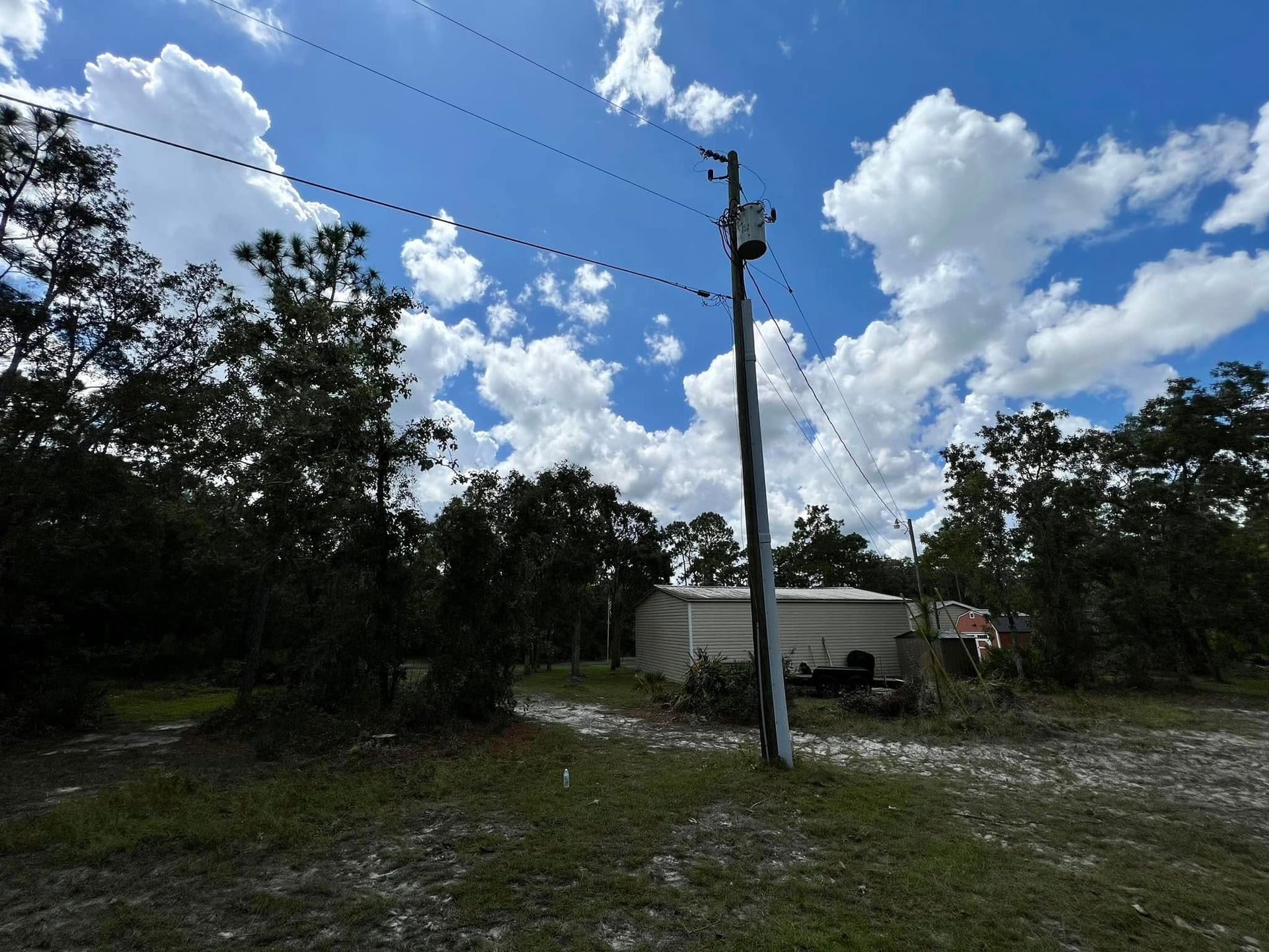 Utility pole with power lines, a structure and trees against a partly cloudy blue sky.
