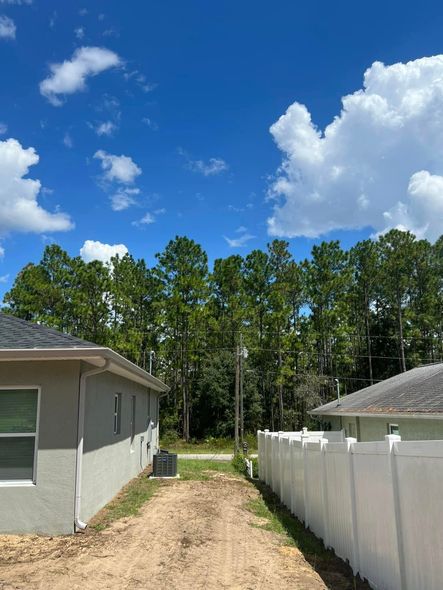 Buildings flank a dirt path leading to a tree line under a blue sky with clouds.