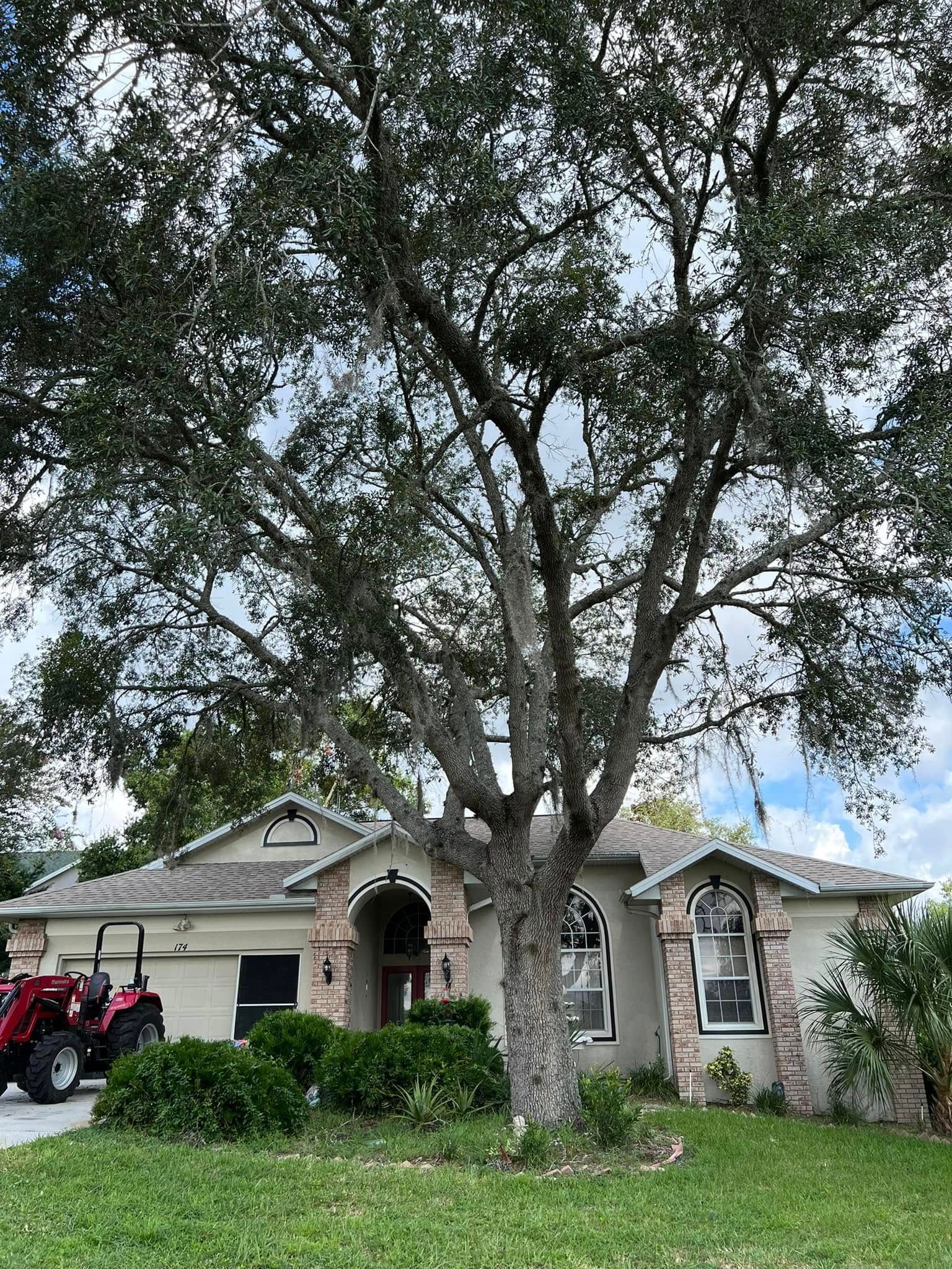 House with a large tree in front. Tractor in the driveway, green grass and bushes, cloudy sky.
