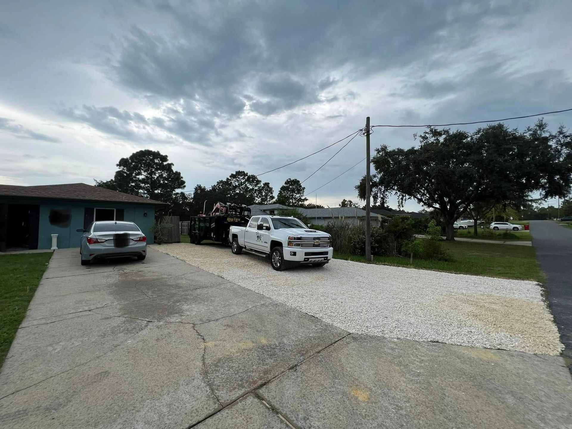 White pickup truck with a trailer parked in a gravel driveway, gray car parked on adjacent concrete. Overcast sky.