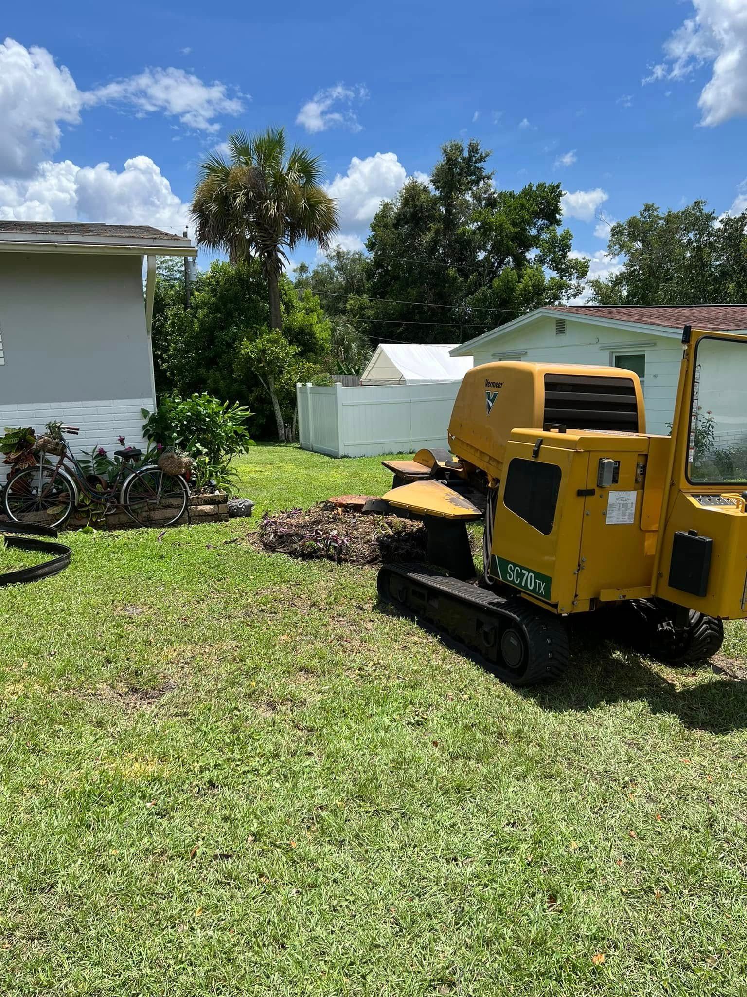 Yellow stump grinder machine in a grassy yard, near a house and trees.