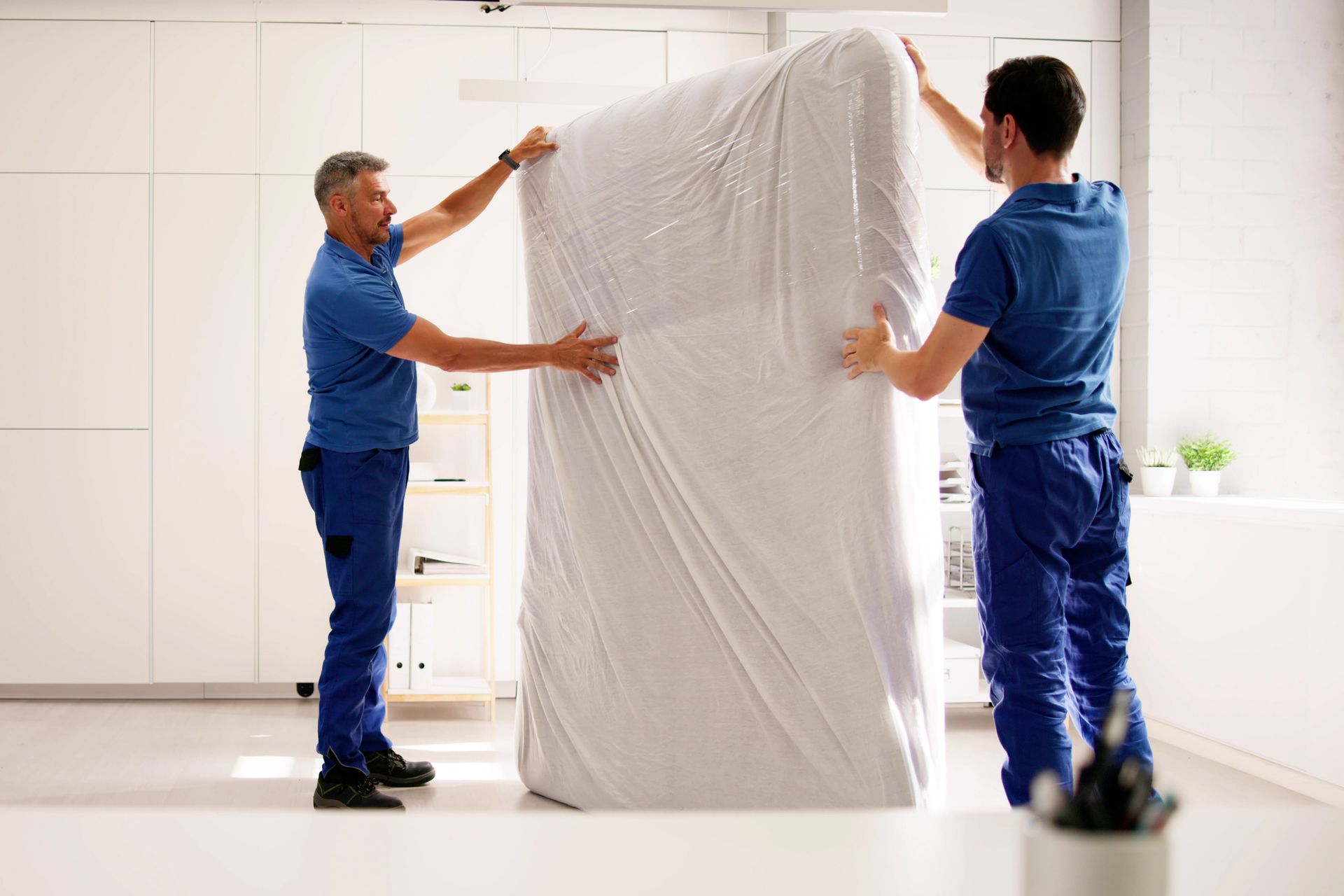 Two men are wrapping a mattress in plastic wrap in a kitchen.