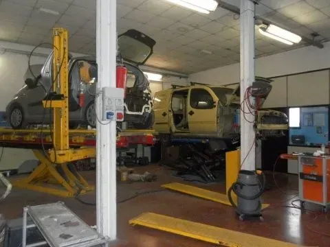 Un'officina meccanica con due auto su ponti sollevatori idraulici, una grigia e una gialla, con vari utensili e attrezzature.