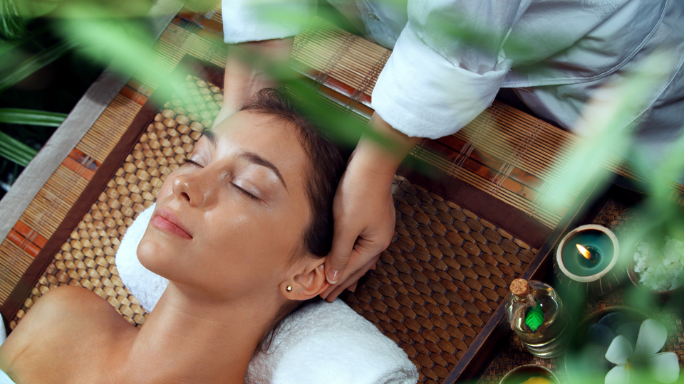 Woman receiving a head massage at a spa; person's hands on her head; lit candles, green accents; relaxed expression.