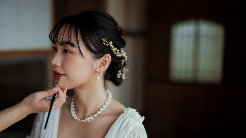 Woman in white dress has makeup applied; pearls, hair adornments, side profile.